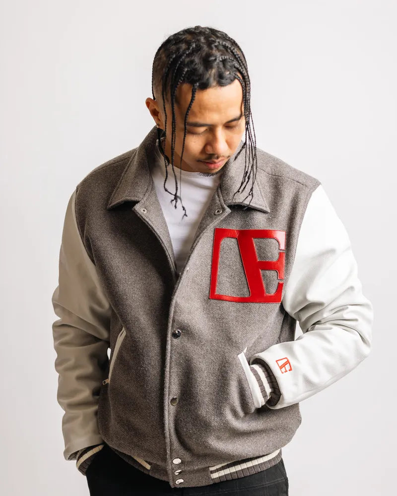 a man is wearing grey letterman jacket while standing